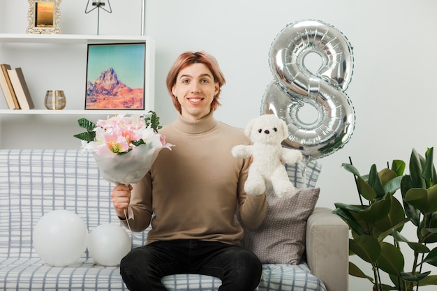 Handsome guy on happy women day holding bouquet with teddy bear sitting on sofa in living room