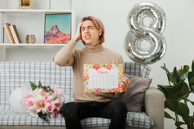 Handsome guy on happy women day holding calendar sitting on sofa in living room