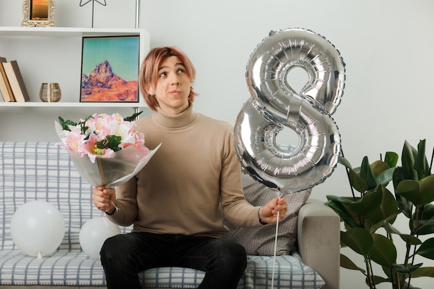 Handsome guy on happy women day holding number eight balloon and bouquet sitting on sofa in living room