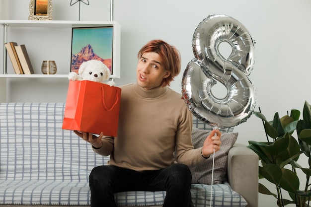 Handsome guy on happy women day holding number eight balloon with gift bag sitting on sofa in living room