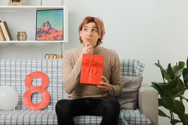 Handsome guy on happy women day holding present sitting on sofa in living room