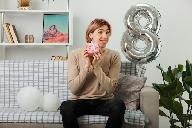 Handsome guy on happy women day holding present sitting on sofa in living room