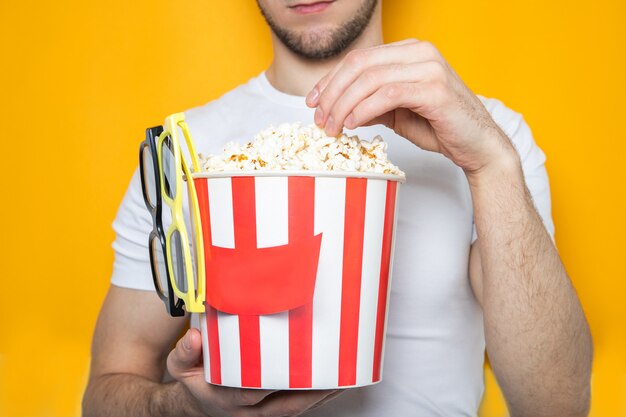 Handsome guy holds popcorn and 3D glasses. Yellow wall. Close-up