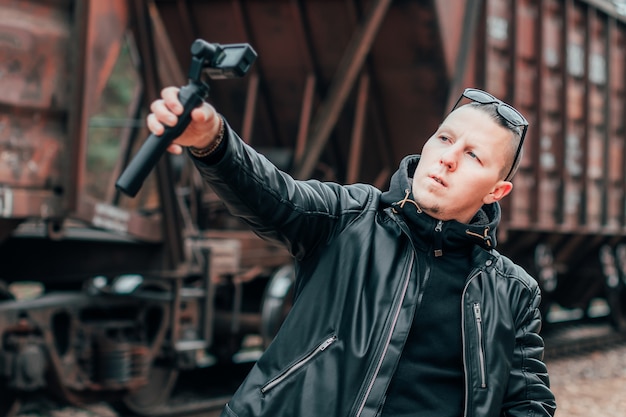 鉄道でジンバルカメラスタビライザーとアクションカメラを使用して自分撮りやストリーミングビデオを作る黒い服とサングラスのハンサムな男