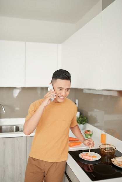 Handsome guy is talking on the mobile phone and smiling while cooking in the kitchen