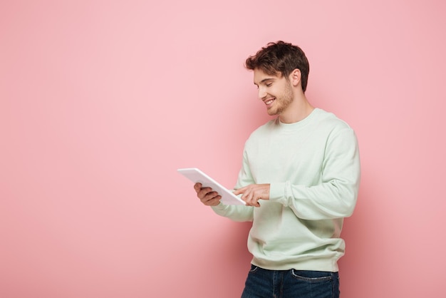 Handsome guy smiling while using digital tablet on pink background