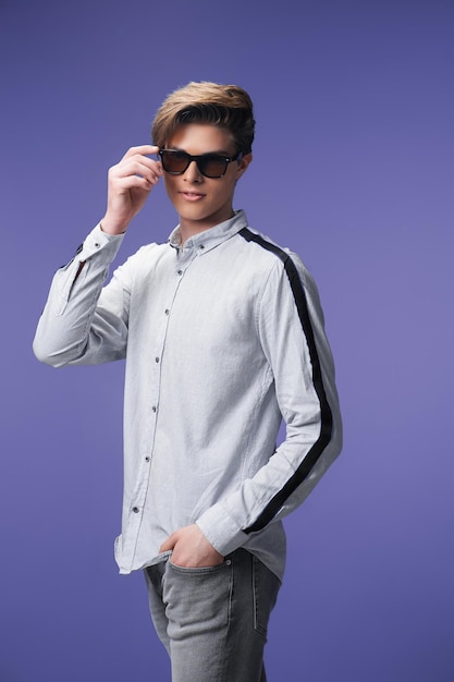 Handsome guy standing in stylish sunglasses holds arm in pocket isolated on purple background