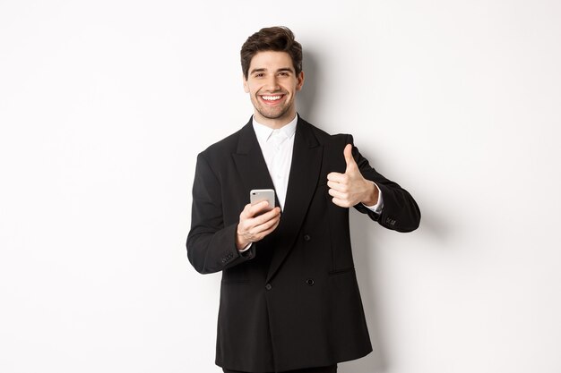 Handsome guy in trendy suit making compliment, showing thumbs-up while using mobile phone, recommending an app or online shop, standing over white background