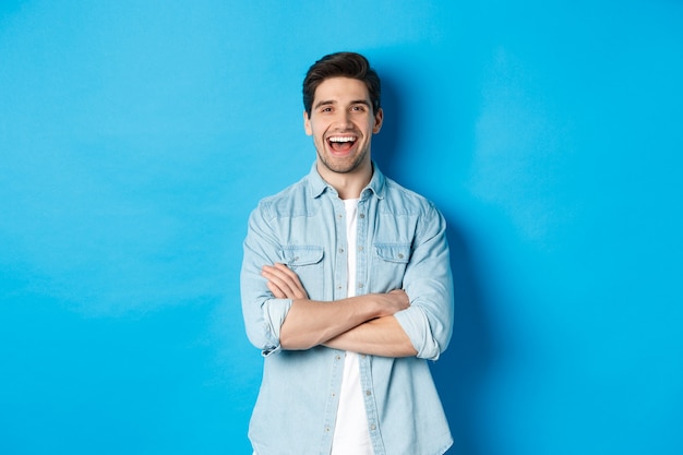 Handsome guy with beard, wearing casual outfit, laughing and looking at something funny, standing against blue wall