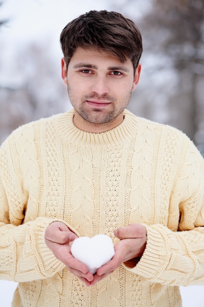 Handsome guy with a knitted sweater holding a heart made of snow in his hands