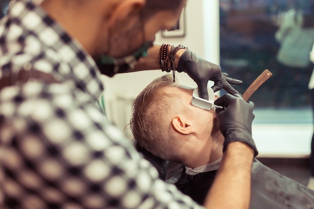 Handsome hairdresser cutting hair of male client. Men hairstylist serving client at vintage barber shop.