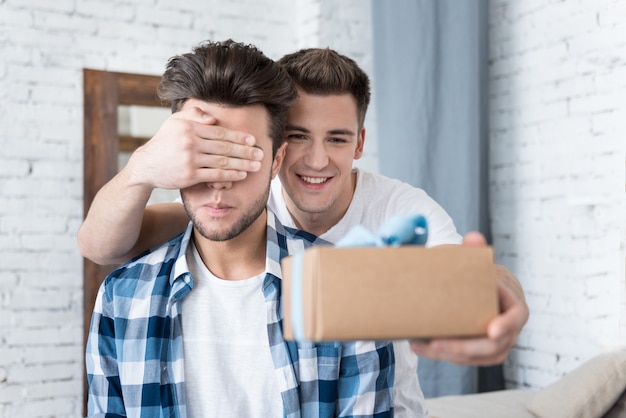 Handsome happy gay man holding a hand over his boyfriends eyes and giving him a present while standing behind him.