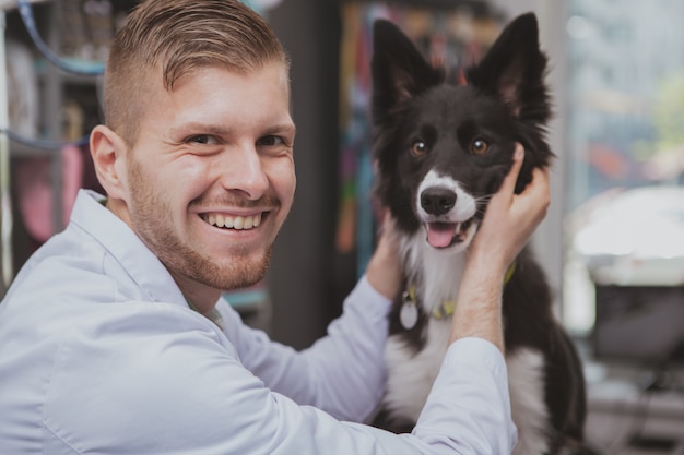 Handsome happy male veterinarian laughing to the camera, petting adorable dog at his clinic