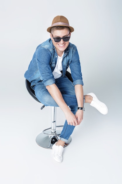 Красивый счастливый человек в джинсовой ткани и солнцезащитных очках сидит на стуле