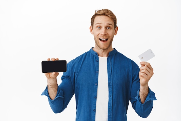 Handsome happy redhead man smiling showing credit card and horizontal smartphone screen recommending application praise bank new feature app standing over white background