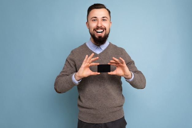 Handsome happy smiling brunette bearded male person wearing grey sweater and blue shirt isolated on background wall holding credit card looking at camera