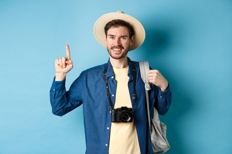 Photo handsome happy tourist in summer hat, holding backpack and camera, pointing finger up at logo, recommending travel agency or place on vacation, blue background.