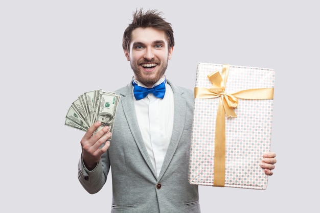 Handsome happy young bearded man in gray suit and blue bow tie standing and holding gift box with yellow bow and many cash dollars with toothy smile face. Indoor, isolated,studio shot, grey background