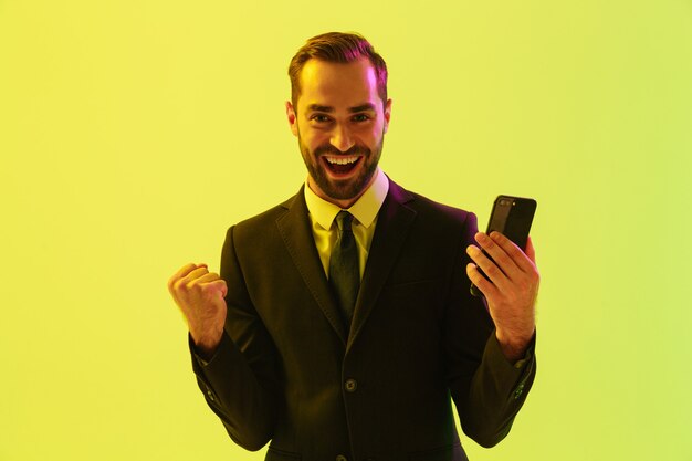 Handsome happy young businessman wearing formal suit standing isolated over yellow wall, holding mobile phone and celebrating success