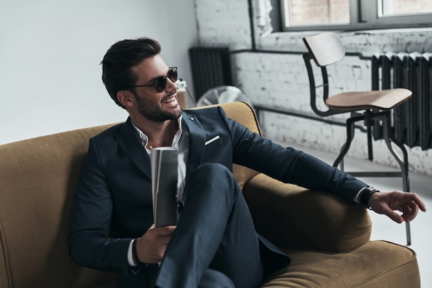 Handsome heartbreaker. Charming young man in full suit looking away and smiling while sitting on the sofa