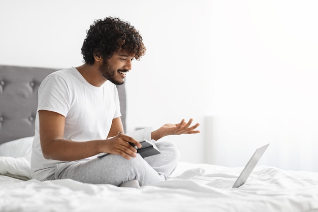 Handsome hindu guy sitting on bed having video conference