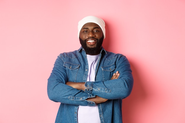 Photo handsome hip hop style black man in beanie and denim shirt, smiling confident, cross arms on chest and staring at camera on pink background.