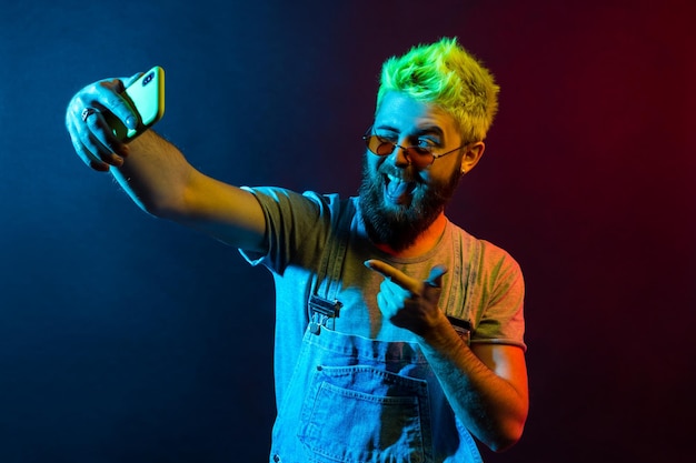 Handsome hipster bearded man wearing t shirt and denim overalls broadcasting live stream points to camera with finger grimacing winking and showing tongue Colorful neon light indoor studio shot