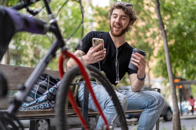 An handsome hipster guy drinks takeaway coffee sitting on a\
bench and making a smartphone video call - gen z people habits\
concept