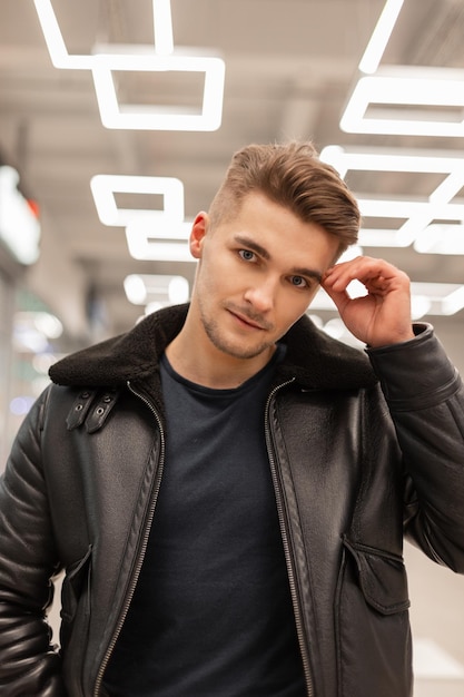 Foto modello di uomo bello hipster con i capelli in maglietta e giacca di pelle nera cammina nel centro commerciale con luci moderne