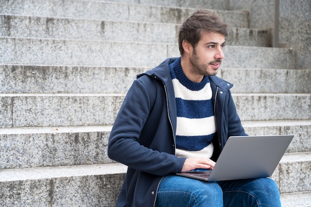 Handsome hipster modern businessman, using laptop in the city, with positive expression.