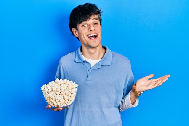 Handsome hipster young man eating popcorn celebrating achievement with happy smile and winner expression with raised hand