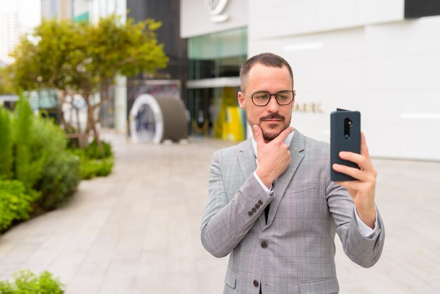 Handsome Hispanic bald bearded businessman taking selfie in the city outdoors