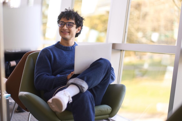 Handsome Hispanic guy student smiles and looks aside browsing websites surfing the net on laptop