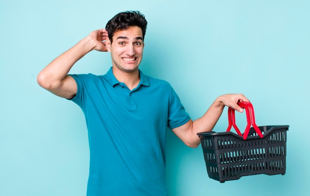 Handsome hispanic man feeling stressed anxious or scared with hands on head shopping basket concept