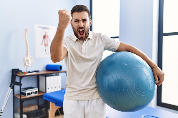 Handsome hispanic man holding pilates ball at rehabilitation clinic annoyed and frustrated shouting with anger yelling crazy with anger and hand raised