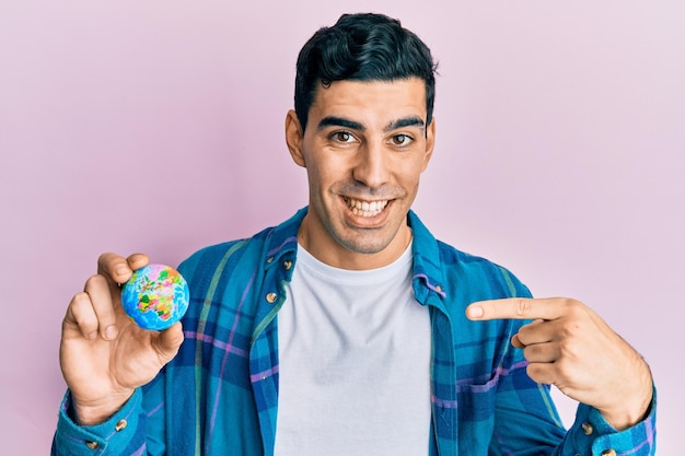 Handsome hispanic man holding small world ball pointing finger to one self smiling happy and proud