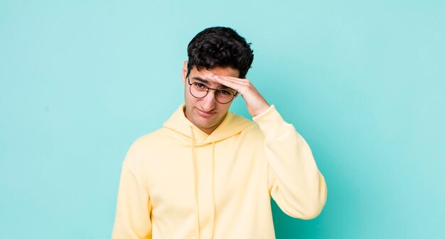 Handsome hispanic man looking bewildered and astonished with hand over forehead looking far away watching or searching