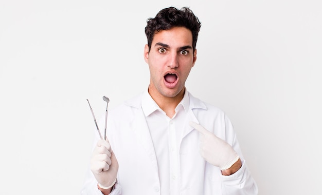 Handsome hispanic man looking shocked and surprised with mouth wide open pointing to self dentist concept