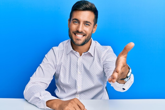 Handsome hispanic man wearing business clothes sitting on the table smiling friendly offering handshake as greeting and welcoming. successful business.