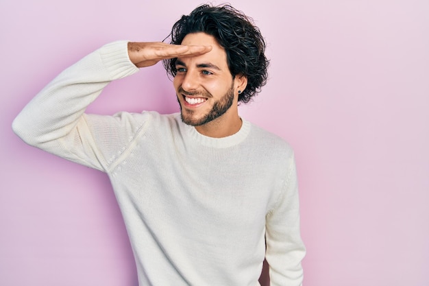 Handsome hispanic man wearing casual white sweater very happy and smiling looking far away with hand over head searching concept