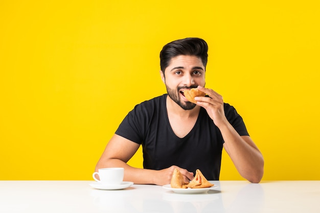 Handsome Indian bearded young man eating Samosa snacks while sitting at white table against yellow background