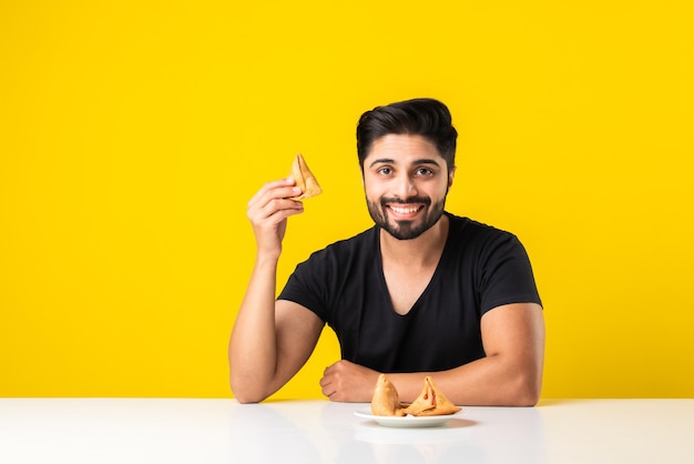 Handsome Indian bearded young man eating Samosa snacks while sitting at white table against yellow background