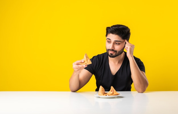 Handsome Indian bearded young man eating Samosa snacks while sitting at white table against yellow background