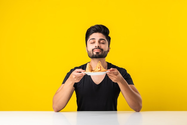 黄色の背景に白いテーブルに座ってサモサスナックを食べるハンサムなインドのひげを生やした若い男