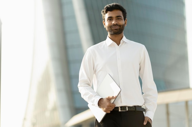 Handsome Indian business man wearing white shirt holding laptop computer looking at camera  