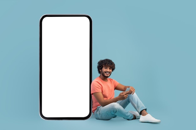 携帯電話を使用して巨大なスマートフォンのそばに座っているハンサムなインド人