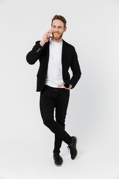 handsome joyful man in jacket laughing and talking on smartphone isolated over white wall