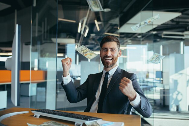 Handsome joyful young businessman sitting at a desk, working in\
a modern office, throwing money over his head. happy smiling man\
throws money in the air