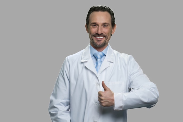 Handsome laboratory assistant giving thumb up gesture. Happy scientist in lab coat on gray background. Symbol of success.