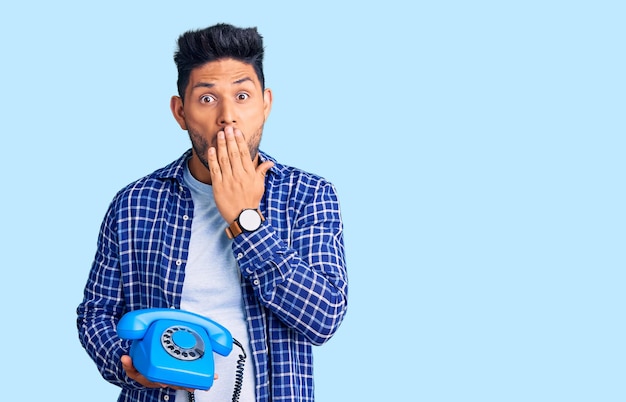 Handsome latin american young man holding vintage telephone covering mouth with hand shocked and afraid for mistake surprised expression